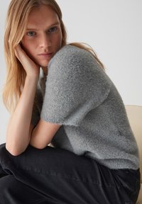 Grijze, korte mouwen, fuzzy gebreid sweater met een relaxte pasvorm. Model zit met gekruiste armen en toont de zachte textuur en subtiele kleurvariaties.