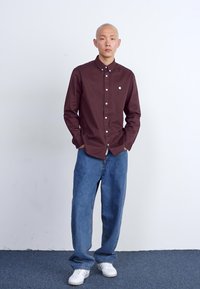Camicia button-up bordeaux con dettagli del logo, abbinata a jeans in denim blu larghi e scarpe da ginnastica bianche su un pavimento texture contro un muro semplice.