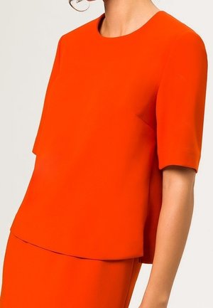 Femme portant un haut orange vif à manches courtes et une jupe assortie sur un fond clair uni.