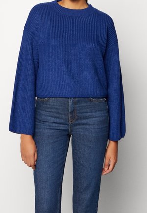 Personne portant un pull en maille épaisse bleu à manches larges, associé à un jean en denim bleu foncé sur un fond uni.