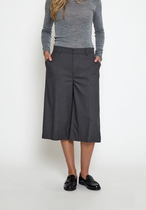 Kvinde iført grå langærmet top, mørkegrå vidde-korte bukser og sorte skinnende loafers, hænderne i lommerne, stående mod hvid baggrund.