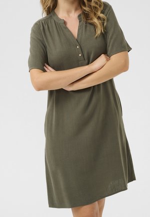 Kobieta ubrana w oliwkowo-zieloną, sięgającą kolan sukienkę z krótkim rękawem i guzikami na piersi, stojąca z założonymi na siebie ramionami na tle gładkiego tła.