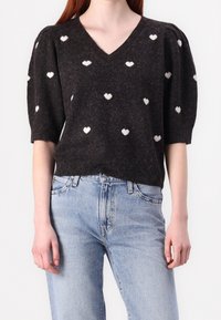 Pull noir en maille avec manches courtes bouffantes, encolure en V et motifs de cœurs blancs partout ; assorti à un jean bleu clair.