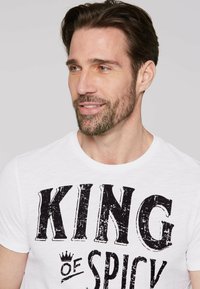 Weißes T-Shirt mit einem auffälligen schwarzen Grafiktext, der "KING of SPICY" mit einer Kronenzeichnung zeigt. Weicher Stoff, Rundhalsausschnitt.
