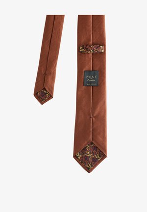 Slips i rostfärgad struktur med blommönstrad foder och etiketten "NEXT Occasion", ligger plant på en vit bakgrund.