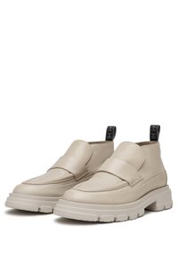 Botas de cuero beige hasta el tobillo con una correa ancha, suela de goma gruesa, parte frontal plana y una lengüeta negra contrastante en el talón.