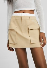 Mini-jupe beige avec deux poches avant, coupe ajustée, et taille blanche élastique. Confectionnée dans un tissu lisse à finition plate.