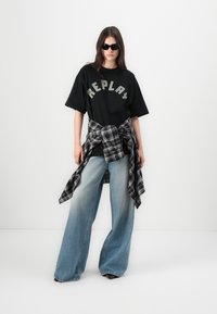 Svart t-skjorte med "REPLAY" i grått, kombinert med lyseblå vide jeans. En svart og grå rutete skjorte knyttet rundt livet. Mørke solbriller.