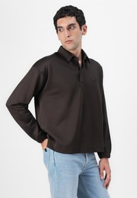 Polo en maille marron à manches longues, fabriqué en tissu lisse, avec un col, une patte de boutonnage et des poignets élastiques. Détail logo discret sur la poitrine.