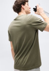 Πράσινο ελιάς βαμβακερό μπλουζάκι με κοντά μανίκια, στρογγυλή λαιμόκοψη και άνετη εφαρμογή, εμφανίζοντας ελαφριά υφή στο ύφασμα, φορεμένο καθημερινά με γκρι παντελόνια.