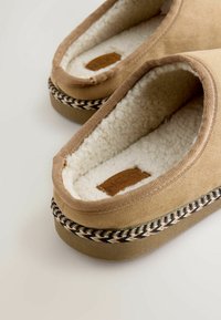 Brown fabric slippers with a soft, cream fleece lining, featuring a woven black and white trim and a small leather label inside.