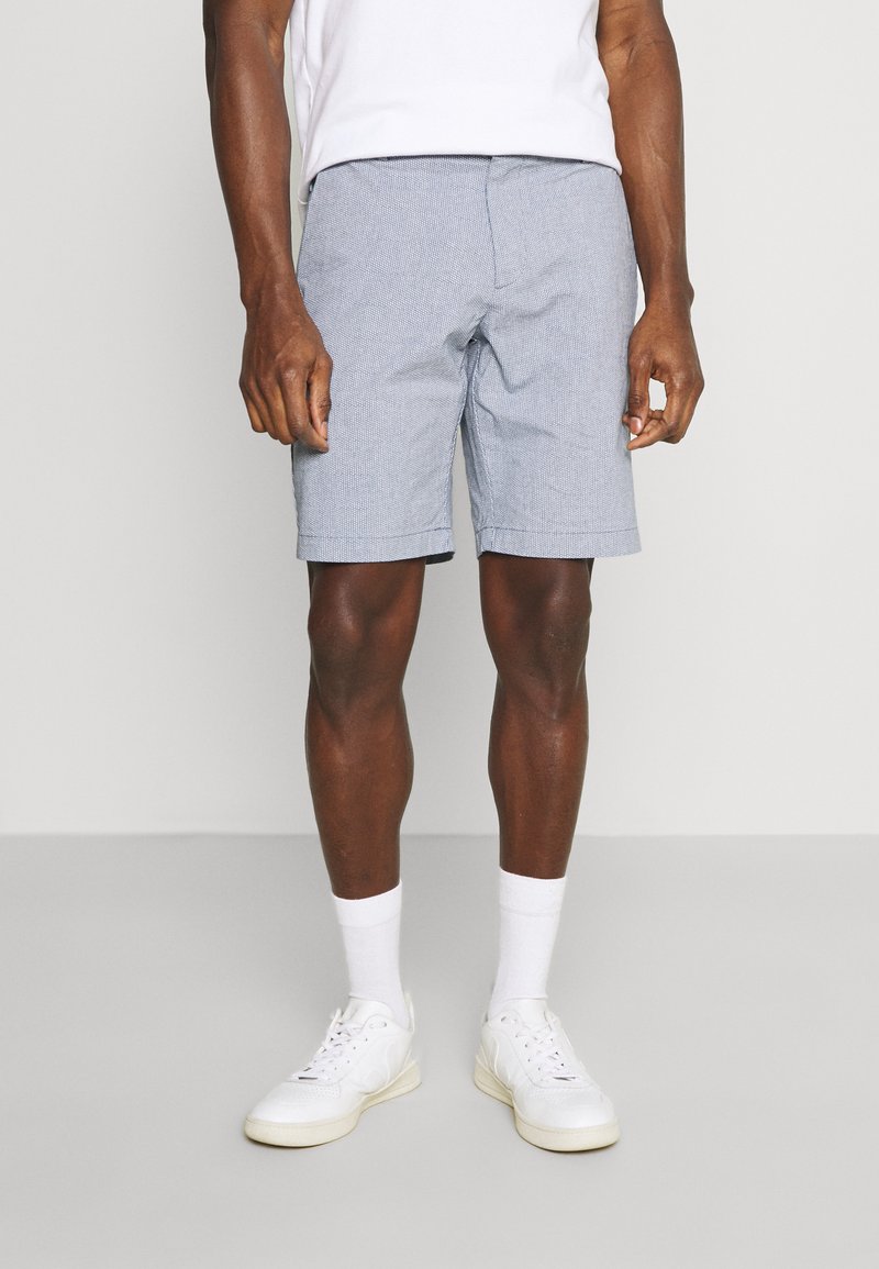 Light blue patterned shorts made from woven fabric. Features side pockets, a flat front, and a straight cut, paired with white sneakers.