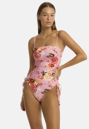 Model poseert in een roze bloemen badpak met verstelbare zijbandjes, met een ontspannen uitstraling en eenvoudige sieraden tegen een effen achtergrond.