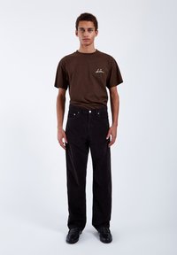 Junger Mann, der ein braunes T-Shirt mit weißem Schriftzug und schwarze Hosen sowie schwarze Schuhe trägt, steht vor einem einfarbigen hellen Hintergrund.