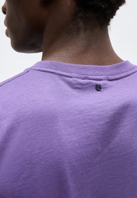 Κοντινό πλάνο του άνω μέρους της πλάτης και του λαιμού ενός ατόμου που φοράει μοβ πουκάμισο με ένα μικρό μαύρο κυκλικό λογότυπο κάτω από το γιακά.