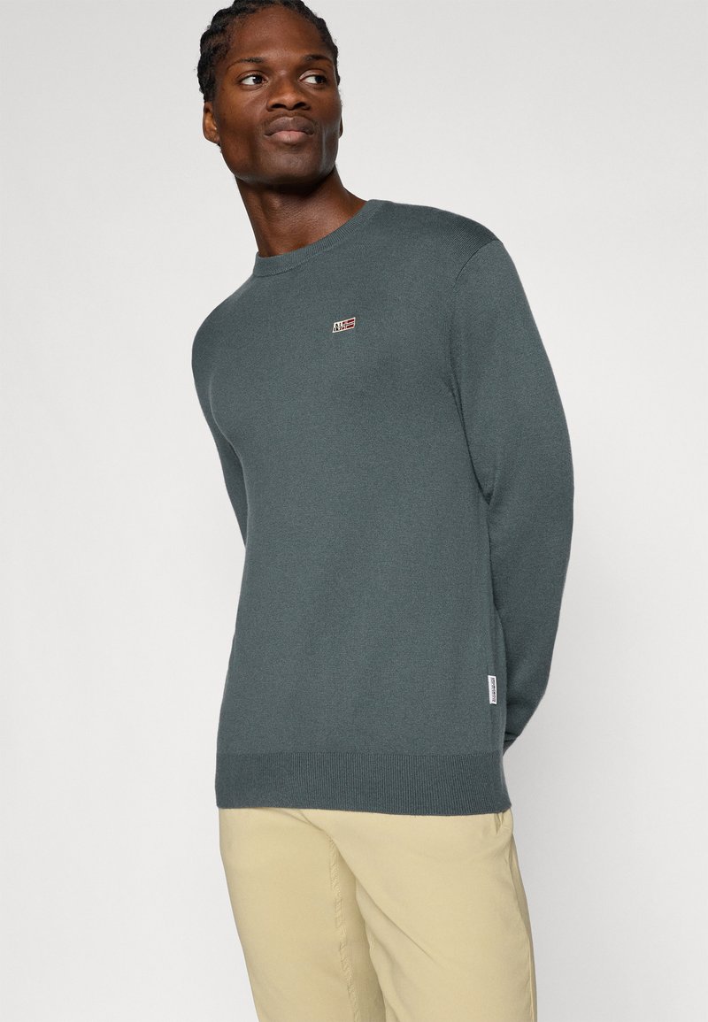 Pull en maille gris avec un col rond, des poignets et un ourlet côtelés, arborant un petit emblème sur la poitrine et une texture lisse.