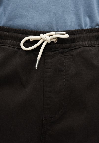 Pantalon noir à taille élastique avec un cordon blanc noué en un nœud sur un fond de tissu de chemise bleue.