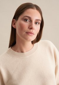 Beige gebreide trui met een ronde ribbelnek en een textuurstof. Het model heeft steil haar, waardoor het ontwerp en de pasvorm van de trui worden getoond.
