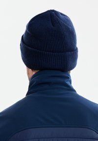 Tummansininen neulottu myssy, jossa on raidallinen rakenne ja käännetty reuna, käytetty yhteensopivan korkeakauluksisen takin kanssa, näkyy takaa.