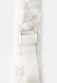 Myk faux fur stropp i hvitt med en teksturert overflate, med en hvit plastspenne og justerbart beige stoffbånd.