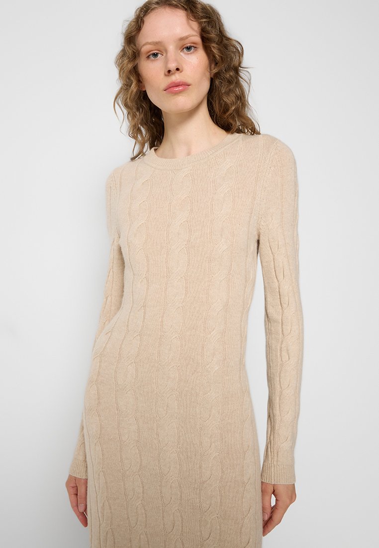 Beiges Strickkleid mit langen Ärmeln, rundem Halsausschnitt und taillierter Form. Weiche Textur mit sichtbaren Zopfmustern und seitlichen Taschen.