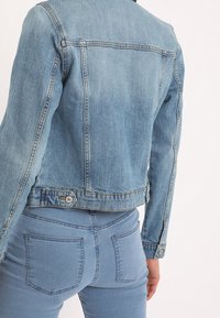 Giacca di jeans azzurro chiaro con design aderente, chiusura con bottoni e cuciture visibili. Indossata con jeans abbinati dello stesso colore.