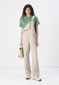 Beige overall met een diagonale klep, groene t-shirt en beige sneakers. Houdt een lichtgekleurde tas vast. Eenvoudig, functioneel ontwerp.