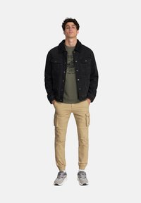 Czarna dżinsowa kurtka z wełnianym kołnierzem, oliwkowa koszulka z grafiką pod spodem, beżowe spodnie cargo i szare sneakersy.