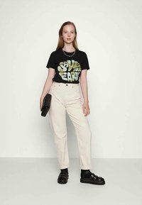 T-shirt preto com gráfico verde "Salve o Nosso Planeta", combinado com calças de cintura alta bege claro e sandálias pretas grossas. Segurando uma clutch preta.