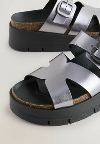 Sandali platform metallici argentati con suole in gomma nera. Presentano ampie cinghie incrociate e un dettaglio con fibbia, mettendo in mostra materiali lisci e texturizzati.