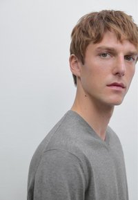 Grauer V-Ausschnitt-Pullover aus weichem Material, mit minimalistischem Design und enganliegender Passform. Der Hintergrund ist in einer hellen neutralen Farbe gehalten.