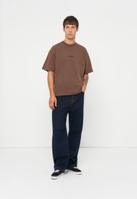 Hnědá oversized bavlněná tričko s krátkými rukávy, spárované s tmavě modrými džínami. Model stojí na bílém pozadí.