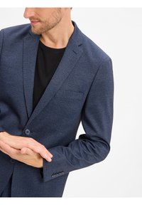 Navy blauwe blazer met een gestructureerd patroon, enkele knoopsluiting, gelaserde revers en een borstzak. Draagt over een zwart T-shirt.