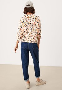 Mehrfarbiges, gemustertes Oberteil in Orange, Schwarz und Creme; 3/4-Ärmel, elastischer Bund, kombiniert mit dunkelblauen Jeans mit umgeschlagenem Saum und hellen Sneakers.