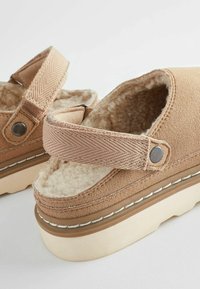 Tan suede platform sandals with a soft, fuzzy interior, adjustable fabric strap, and round metal accents on the sides.