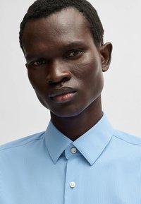 Camicia a righe blu chiaro con colletto, dotata di una chiusura a bottoni. Il tessuto ha una texture liscia e una leggera lucentezza.