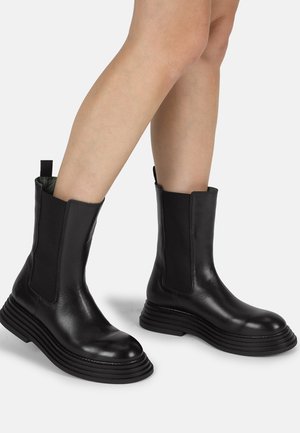 Botas de tobillo de cuero negro con paneles elásticos laterales, suelas gruesas y estriadas, y una tira en la parte trasera para facilitar el calzado. Textura suave.