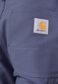 Tejido azul con un acabado texturizado, que presenta un bolsillo visible y un parche del logo de Carhartt en blanco y amarillo. Detalle de costura evidente.