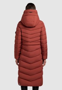 Long puffer coat in rust red with chevron quilting, attached hood, and long sleeves. Smooth texture and slim fit design.