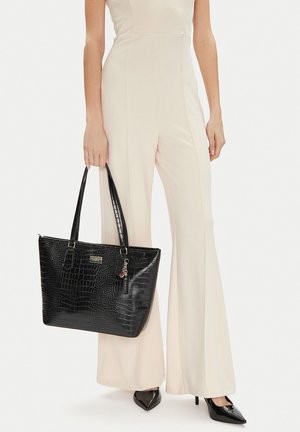 Žena v krémovém kostýmku s kalhotami drží velkou černou kabelku s krokodýlím vzorem, dvojitými uchy a ozdobným přívěskem.