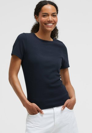 Smiling woman with curly hair wears a fitted short-sleeve navy shirt and white pants with hands in pockets against a plain background.