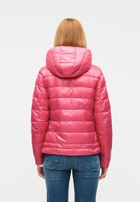 Γυναίκα με ίσια  ξανθά μαλλιά, που φοράει ένα φωτεινό ροζ ντυμένο με επένδυση μπουφάν με κουκούλα και μπλε τζιν, εμφανίζεται από πίσω σε λευκό φόντο.