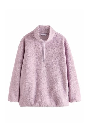 Personne portant un pull rose duveteux avec une fermeture éclair et un pantalon noir à jambes larges avec un motif zèbre subtil, debout devant un fond uni.