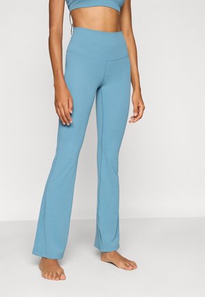 Žena stojící naboso na neutrálním pozadí, oblečená ve světle modrých kalhotách na jógu s vysokým pasem a rozšířenými nohavicemi a odpovídajícím krátkém topu.