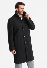 Abrigo de lana negro con un cuello alto, frente de botones ocultos y bolsillos laterales. Presenta un corte recto y una textura suave.