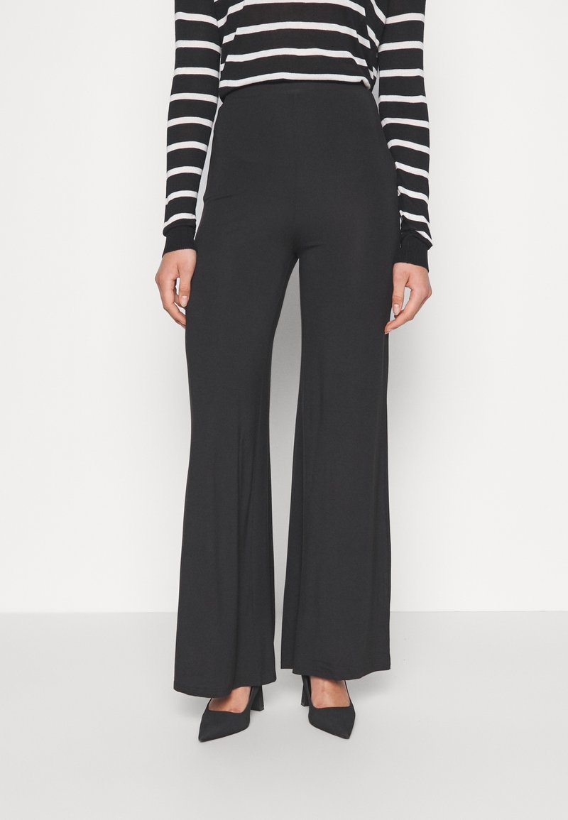Czarne spodnie z wysokim stanem wykonane z gładkiego materiału, z rozszerzanym krojem nogawki. W zestawie z czarną i białą, paskowaną bluzką z długim rękawem.