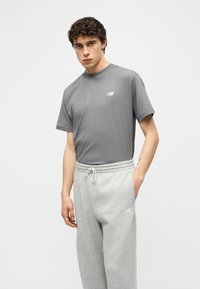 Νέος άνδρας που φοράει γκρι μπλουζάκι New Balance και ανοιχτό γκρι φόρμα, στέκεται με το ένα χέρι στην τσέπη μπροστά σε απλό φόντο.