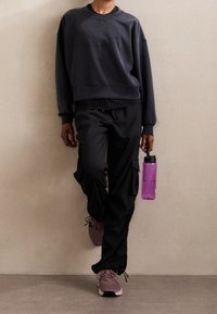 Sudadera gris de manga larga, pantalones cargo negros y zapatillas deportivas rosas. Sosteniendo una botella de agua morada, de pie contra una pared neutra.