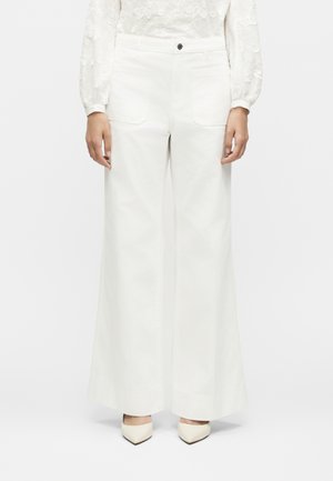 Kvinde iført hvide, vidde bukser med høj talje og forlommer samt hvide spidse hæle, stående mod en ensfarvet baggrund.