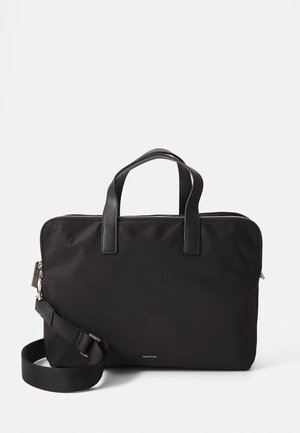Sac d'ordinateur portable noir en nylon avec des accents en cuir. Il dispose de deux poignées supérieures, d'une bandoulière amovible et d'une fermeture à glissière. Branding minimal.